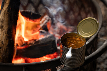 A can of food heating up next to a fire. The fire is raging and the can is silver.