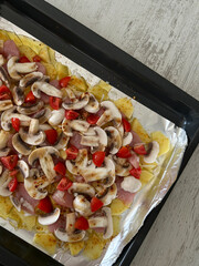 Baking tray with raw sliced potatoes, mushrooms, tomatoes, and meat on foil, prepared for cooking. Used in food, cooking, and recipe contexts.