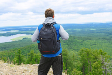 A man in a blue tank top and gray turtleneck stands on a mountain top with a backpack.