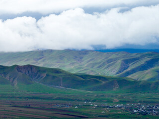 Close-up view of lush green, rounded foothills rising above a distant plain and village, topped by a heavy layer of dramatic white and blue clouds