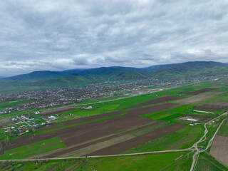 High aerial view of a village situated between geometrically patterned fields and the base of prominent green, rolling mountains under a moody, cloudy sky