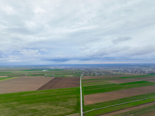 Expansive aerial view of a straight road dividing vast sections of tilled brown earth and vibrant green crops, extending towards a distant city under an overcast sky
