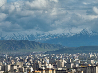 Close-up view of Bishkek city's dense residential buildings set against the dramatic backdrop of green hills and snow-capped mountains under dark, cloudy sky