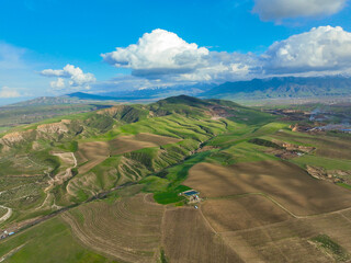 Wide aerial view of lush green rolling hills and geometrically patterned farmland, with the distant snow-capped mountains under a bright, cloudy sky
