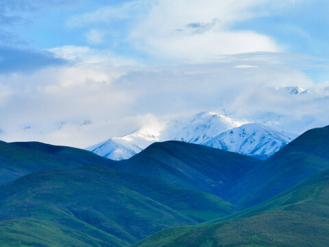 Dramatic landscape composition of deep blue-green, rolling hills in the foreground contrasting with bright, sunlit snow-capped mountains beneath a cloudy sky - Powered by Adobe