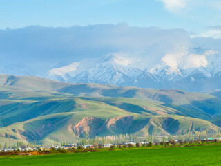Fototapeta premium Close-up landscape of bright green, rolling foothills and a village at their base, with towering snow-capped peaks partially obscured by clouds above