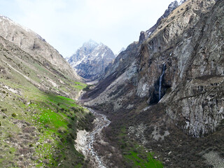 Wide view of a deep mountain valley with bright green sections, a central riverbed, and a small waterfall flowing from the steep right cliff