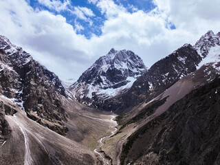 Aerial view centered on a prominent snow-covered mountain peak, flanked by steep slopes, with a winding riverbed leading into the valley floor