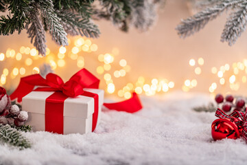 White gift box with red ribbon and ornaments under snowy branches, set against warm holiday lights. Winter Christmas Holiday Decoration Background