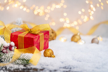 Festive red gift box wrapped in gold ribbon rests on snow with frosted pine, berries, and glowing lights in the background, creating a cozy holiday atmosphere. Ideal for holiday greeting cards,