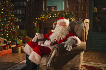 Merry Christmas. Happy Santa Claus sitting in chair in festive living room with fireplace...