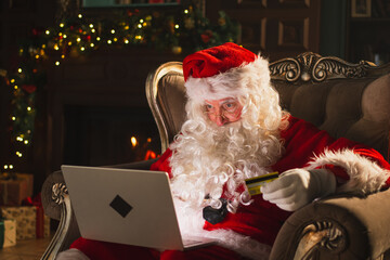 Merry Christmas. Santa Claus shopping online holding laptop paying with gold credit card for...