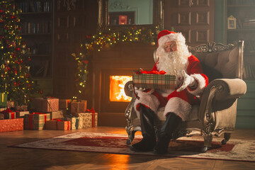 Merry Christmas. Santa Claus sitting in chair packing wrapping gift box in festive living room with...