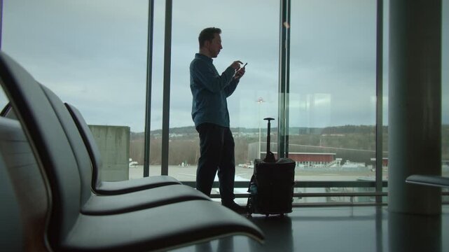 A traveler stands at the airport window gazing out while holding their luggage patiently in hand