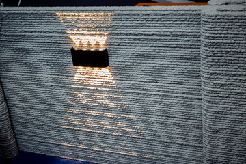 Textured 3D printed wall of layered concrete or plaster, illuminated by a modern up/down wall light. Showcases innovative architectural design and construction.