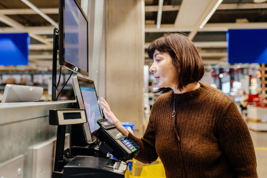 Mature woman over 55 taking printed receipt at self service checkout in furniture store. Senior person completing payment independently, demonstrating confidence and digital skills.