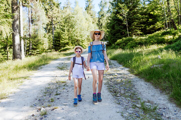 Fototapeta premium Two sisters hikers with backpacks, hats and sunglasses walking together along a sunny forest trail, summer outdoor adventure, family bonding and active lifestyle in nature scene