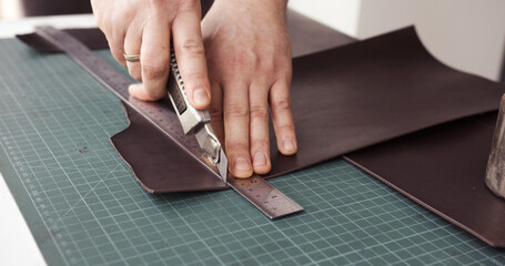 Leather craftsman cutting brown leather sheet with utility knife and metal ruler
