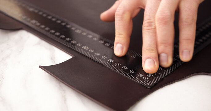 Hand measuring brown leather sheet with metal ruler on white table during crafting process