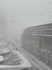 Large building and snowy street with cars and bus during heavy snowfall. Used in architecture, transport, and climate materials.