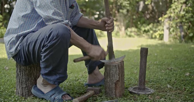 A close-up view of a man diligently sharpening an axe using a hammer and chisel, seated on a tree stump in an outdoor setting. This video features authentic tool sounds.