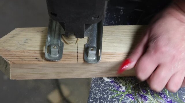 A close-up shot captures a woman meticulously using a jigsaw to cut through a piece of wood in her home, her red nails a bright contrast to the purple flower decoration of the table.