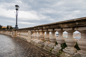A weathered stone balustrade and lamppost line a curved cobblestone path, offering a distant view of a city harbor under a cloudy sky.