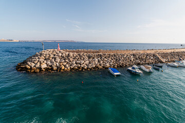 A rocky breakwater with a striped lighthouse extends into the vibrant blue sea, protecting several moored boats under a clear sky.