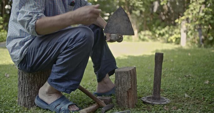 An elderly man meticulously sharpens an old axe using traditional hand tools while sitting on a tree stump in a lush green garden. This video captures the authentic sounds of his focused labor.