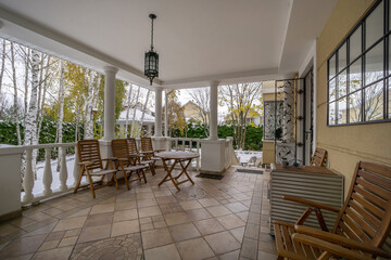 A peaceful porch features wooden chairs and a small table, offering a cozy spot to relax. Bare trees surround the area, indicating the chill of winter.