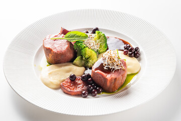 A gourmet plate of fine-dining meat medallions with mashed potatoes, broccoli, and a berry-based sauce, garnished with sprouts. The dish is professionally styled on a white ridged plate.