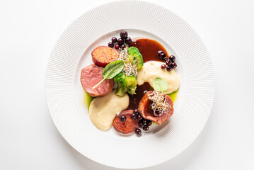 A gourmet plate of fine-dining meat medallions with mashed potatoes, broccoli, and a berry-based sauce, garnished with sprouts. The dish is professionally styled on a white ridged plate.