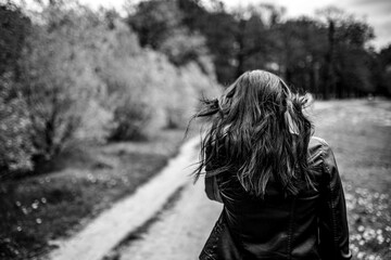 A woman in a leather jacket with long dark hair blowing walks away down a park path. The monochrome image emphasizes the windblown texture, volume, and natural movement of her dark hair.