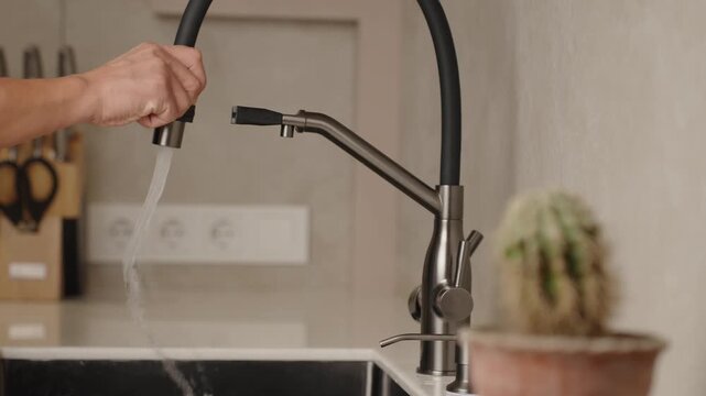 Unknown person expertly washes the sink with sleek modern faucet in bright kitchen. Unrecognized man showcasing functions of flexible hosepipe of trendy faucet.