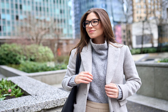 Young happy elegant brunette woman wearing coat and glasses standing on big city street. Smiling business lady walking outdoors looking away enjoying fall time in downtown.