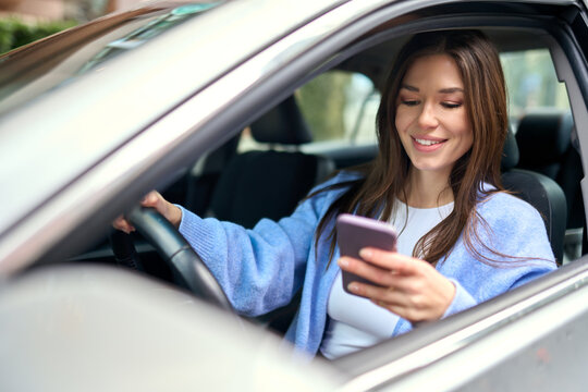 Young happy woman driver sitting inside car holding smartphone using gps navigation map app on mobile phone looking at cellphone chacking traffic on city roads. Driving applications concept.