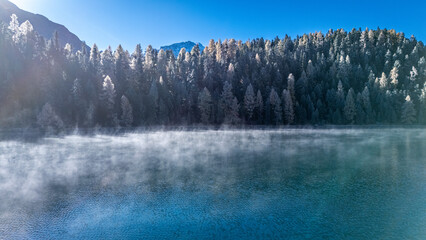 Drohnenaufnahme eines mystischen Bergsees im Engadiner Herbst: Nebelschwaden schweben über...