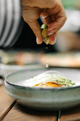 Close-up of a hand squeezing lime over a dish with microgreens and vegetables. Fresh citrus juice drops falling on food. Natural light and soft focus food photography.
