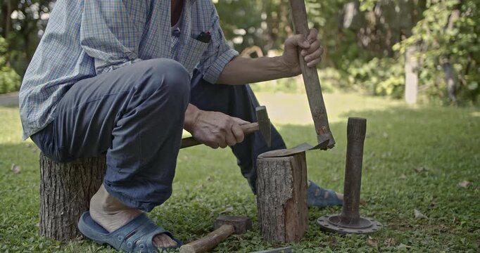 A person sits on a tree stump, carefully hammering a hand tool into a piece of wood, performing manual labor outdoors. This video includes crisp sounds of work.