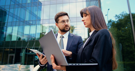 Two business professionals in formal attire standing outdoors, collaborating on remote work using laptop and tablet in a modern urban environment