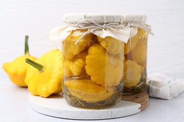 Pickled pattypan squashes in jars and fresh vegetables on white table, closeup