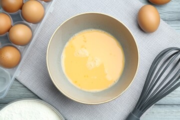 Beaten eggs in bowl, whisk and flour on table, flat lay