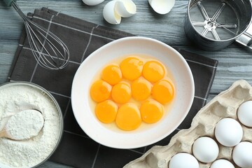 Raw eggs, yolks in bowl, flour, sifter and whisk on wooden table, flat lay