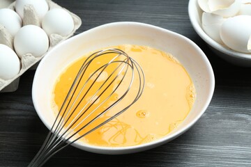 Beaten eggs in bowl and whisk on dark wooden table, closeup