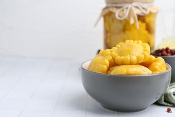 Pickled pattypan squashes in bowl on white tiled table, closeup. Space for text