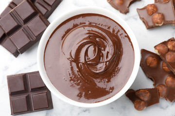 Bowl with melted chocolate and pieces on white marble table, flat lay