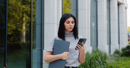 Business woman wearing suit using smartphone walking in big city on street outdoor. Female professional executive typing on cellular phone, online mobile technology business app while holding laptop.