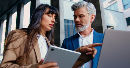 Close up of two business people, a man and a woman in formal attire, are standing outside a modern office building, discussing work-related topics using a digital tablet and a laptop