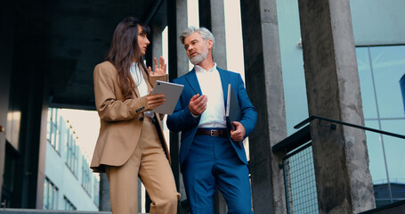Young businesswoman and older businessman in formal suits walking down stairs after work, using laptop and tablet, discussing work-related topics