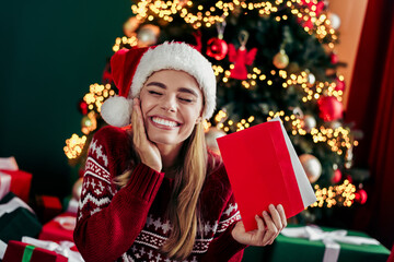Funny festive moment a smiling woman in a Santa hat opens a red gift beside a glowing Christmas tree with warm lights and wrapped presents creating a cozy home holiday scene
